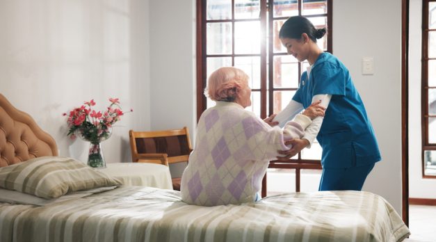 Senior woman, caregiver and help on bed with care, assisted living and support in nursing home. Elderly patient, nurse and mobility aid in bedroom for friendly service, healthcare and consulting