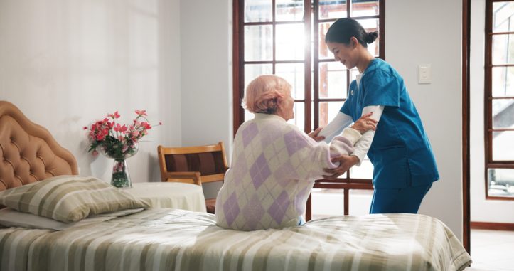 Senior woman, caregiver and help on bed with care, assisted living and support in nursing home. Elderly patient, nurse and mobility aid in bedroom for friendly service, healthcare and consulting